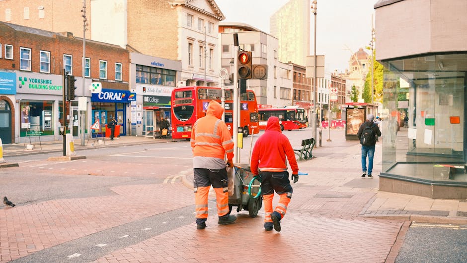 Two workers dressed in high-visibility orange and red safety clothing are walking along a busy urban street, pushing a small hand trolley loaded with debris and rubbish. They are positioned in the foreground, moving away from the camera, with one worker holding the trolley handle while the other guides it. The street is lined with various commercial storefronts, including a building society and a pharmacy, with signs visible in the background. Behind them, there is a red double-decker bus stopped at a traffic light, which is red, and other buses and pedestrians are seen in the distance. The sidewalk is paved with red bricks and concrete, and there are street benches near the workers, with a few pedestrians walking or waiting at bus stops. To the right, a storefront with large glass windows reflects part of the street scene. The lighting appears natural, suggesting daytime, and the overall scene depicts a typical city environment where rubbish removal or on-site clearance may be relevant, as performed by local waste management services like Rubbish Clearance Tooting.