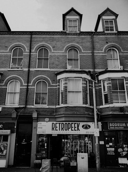 A three-story brick building with a commercial ground floor featuring a sign for 'Retropeek House Clearances' in bold, black lettering on a white background. The building has large, arched windows on the second floor with white framed glass panes, and smaller rectangular dormer windows on the attic level with peaked roofs. The upper floors are constructed from red brick with some decorative white brickwork accents, and the windows have white sills. In front of the building, there are a few chairs and a small table, indicating outdoor seating or waiting area. To the right, part of another storefront with a sign mentioning 'Bodrum,' a Turkish restaurant or shop, is visible. The overall scene is in black and white, capturing a street-level view of an independent waste clearance service operating within a commercial property, likely involved in rubbish removal or disposal handled as part of private on-site clearance services integrated into the urban environment.