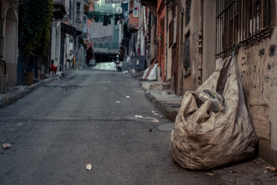 Two workers dressed in high-visibility orange and red safety clothing are walking along a busy urban street, pushing a small hand trolley loaded with debris and rubbish. They are positioned in the foreground, moving away from the camera, with one worker holding the trolley handle while the other guides it. The street is lined with various commercial storefronts, including a building society and a pharmacy, with signs visible in the background. Behind them, there is a red double-decker bus stopped at a traffic light, which is red, and other buses and pedestrians are seen in the distance. The sidewalk is paved with red bricks and concrete, and there are street benches near the workers, with a few pedestrians walking or waiting at bus stops. To the right, a storefront with large glass windows reflects part of the street scene. The lighting appears natural, suggesting daytime, and the overall scene depicts a typical city environment where rubbish removal or on-site clearance may be relevant, as performed by local waste management services like Rubbish Clearance Tooting.