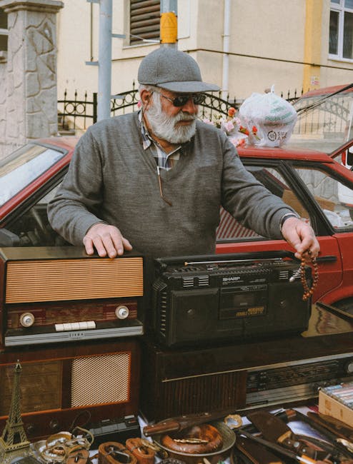 A middle-aged man with a white beard and sunglasses, wearing a gray flat cap and a light gray zip-up sweater over a checkered shirt, stands behind a red vehicle on a city street. He is holding a vintage portable radio with both hands, positioning it among various other objects. In the foreground on a table or surface, there are multiple antique items including a small Eiffel Tower figurine, leather belts, and a wooden box. Behind him, a plastic bag with some items inside is placed on the vehicle's roof, which is parked on a paved sidewalk near a building with cream-colored walls, a black metal fence, and a window with open shutters. The overall scene appears to be part of an outdoor market or second-hand sale, with the man engaged in arranging or displaying items for sale, possibly related to private or alternative waste handling and rubbish collection services like those offered by Rubbish Clearance Tooting.