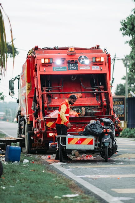 A waste management worker dressed in a high-visibility yellow and orange reflective vest is operating a large red rubbish collection truck on a paved roadside area. The truck has a sturdy, metallic texture with visible hydraulic mechanisms on its rear, used for compacting and loading waste. The scene includes the worker standing close to the truck, slightly leaning forward while handling a black plastic bag filled with waste, which is placed on a small metal platform attached to the vehicle. The surrounding environment features a road with white edge lines, a grassy verge with a small blue container on the ground, and some scattered trash around the worker’s feet. In the background, there are trees, utility poles, and a distant storefront with signage, under a daylight sky that appears overcast. The overall scene suggests an on-site waste collection operation, possibly by a private waste handling service like Rubbish Clearance Tooting, emphasizing independent rubbish removal activities in a residential or commercial area.
