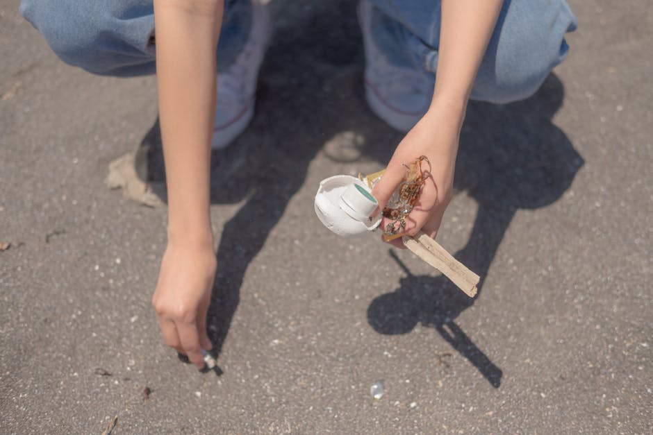 A person's hands are visible in the image, with one hand reaching towards the ground and the other holding a small bundle of debris consisting of a broken white ceramic mug with visible green interior, tangled with dried plant material, on a light wooden stick. The individual is dressed in blue jeans and grey shoes with red accents. The scene takes place on a paved surface, possibly a street or driveway, with little visible background other than the concrete. The lighting suggests natural daylight, and the image captures a moment of cleaning or waste collection, reflecting a focus on rubbish removal or environmental cleanup activities. The detailed textures of the debris and the ground are clearly visible, emphasizing the act of picking up and disposing of waste, a key aspect of services like those offered by Rubbish Clearance Tooting.